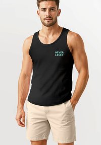 Mann, der selbstbewusst in einem schwarzen Tank-Top mit dem Text "NEVER LESS®" und hellbeigen Shorts vor einem neutralen Hintergrund posiert.