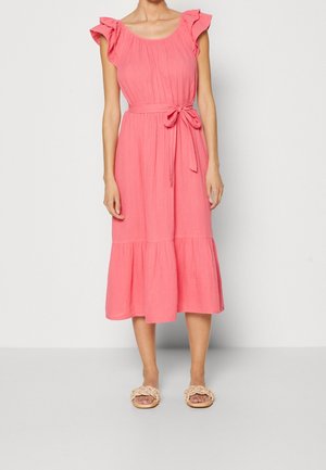 Vrouw die een mouwloze koraalrode midi-jurk draagt met gerimpelde schouders, een geknoopte tailleband en beige gevlochten sandalen.