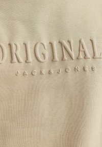 Beidzis džemperis ar teksturētu, izvirzītu uzrakstu "ORIGINAL JACK & JONES". Audums izskatās mīksts ar gludu tekstūru.
