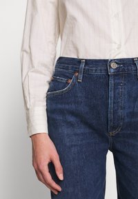 Personne portant un jean en denim bleu foncé et une chemise blanche à manches longues avec de fines rayures verticales, une main détendue le long du corps.