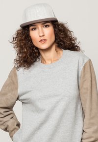 Grijze sweatshirt met beige suède mouwen; een lichtgrijze pet met een flatte rand. Het model heeft krullend haar en een neutrale uitdrukking.