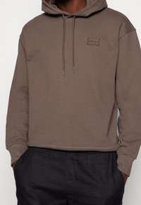 Felpa marrone con tasca a marsupio, cappuccio con cordino e patch logo cucito. Materiale in cotone morbido con vestibilità comoda e polsini a coste.