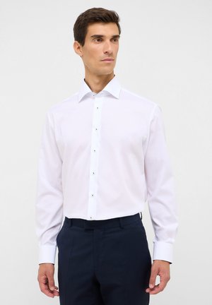Weißes Hemd mit langen Ärmeln, knöpfbarem Frontbereich und kontrastierenden Knöpfen. Glattes Material, strukturierter Kragen, getragen mit dunklen Hosen.