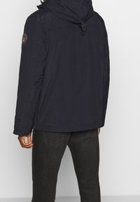 Giacca imbottita blu navy con cappuccio, dotata di tasche con cerniera e toppa con logo sulla manica, tessuto strutturato con finitura impermeabile.