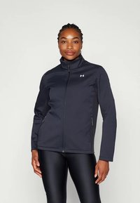 Mujer con el cabello trenzado que lleva una chaqueta negra de cremallera con cuello alzado y leggings negros de apariencia de cuero, de pie contra un fondo liso.
