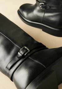 Juodos odinės iki kelio aukščio botai su lygiu paviršiumi, turintys sagties dirželio detalę ir storą, tekstūruotą pado dalį.