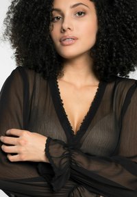 Schwarze transparente Bluse mit tiefem V-Ausschnitt und floralem Spitzenbesatz. Verfügt über geraffte Puffärmel und einen leichten, strukturierten Stoff.