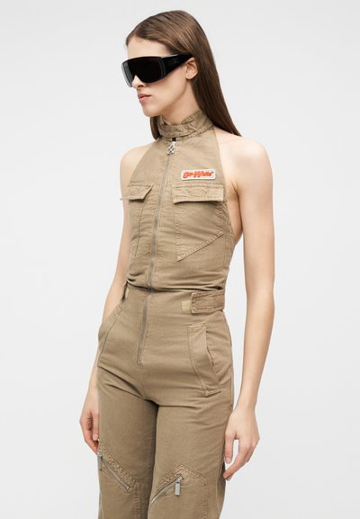 Γυναίκα που φοράει μπεζ ολόσωμη φόρμα χωρίς μανίκια με φερμουάρ μπροστά και μαύρα μεγάλα γυαλιά ηλίου, κοιτάζοντας προς τα αριστερά μπροστά σε απλό φόντο.