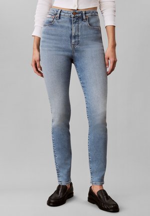 Personne portant un jean skinny bleu clair, un haut court blanc et des mocassins en cuir noirs, debout devant un fond gris uni.