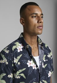 Camisa de manga corta con una base azul marino oscuro, decorada con hojas verdes y patrones florales blancos, hecha de un tejido ligero y con un diseño de cuello.