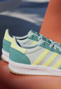 Ein Paar türkise, weiße und gelbe Adidas-Sneaker mit Cloudfoam-Sohlen auf einer orangenen Oberfläche im natürlichen Licht.