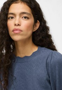 Mujer con cabello oscuro rizado que lleva un suéter azul texturizado, mirando directamente al frente con una expresión neutral contra un fondo simple.