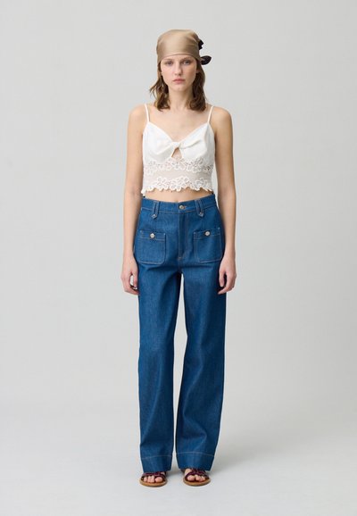 Top court en dentelle blanche avec détails et un noeud à l'avant, associé à un pantalon en denim bleu ample avec deux poches avant. Sandales marron.