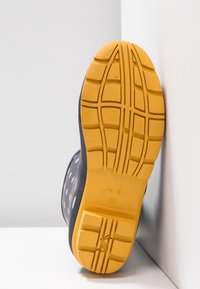 Tumman sininen vedenpitävä saappaat, joissa on valkoisia pilkkuja. Saappaissa on teksturoitu keltainen kumipohja, jossa on kuvioitu pinta ja logo sivussa.