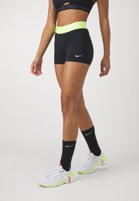 Schwarze Nike Pro Shorts mit einem neonfarbenen gelben Bund, der das Nike-Logo trägt. Kombiniert mit schwarzen Socken und weißen Sneakers mit pinken und grünen Akzenten.