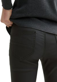 Schwarze, eng anliegende Hose aus strukturiertem Stoff mit einer aufgesetzten Gesäßtasche und kontrastierenden Nahtdetails. Darüber ist ein dunkler Pullover zu sehen.