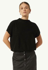 Schwarzer Strickpullover mit kurzen Ärmeln, hohem Kragen und verstreuten silbernen Akzenten. Kombiniert mit einem schwarzen Rock mit Knopfdetails.