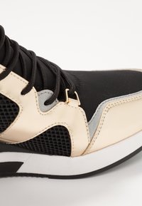 Czarno-złoty but sportowy z siateczkową cholewką, gładkimi złotymi i szarymi detalami oraz białą i czarną podeszwą, o zaokrąglonym kształcie i teksturowanym wzorze.