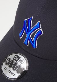 Căciulă de baseball ajustabilă, de culoare albastru închis, cu logo-ul NY brodat în albastru deschis. Prezintă un material texturat și un etichetă de marcă circulară pe față.