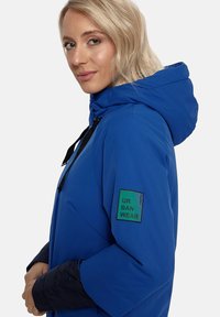 Zils ūdensizturīgs mētelis ar kapuci, ar melniem akcentiem uz aprocēm un zaļu logo uz pleca. Gluda tekstūra.