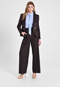 Braun maßgeschneiderte zweireihige Blazer mit goldenen Knöpfen, hellblauem Streifenhemd, hochtaillierten braunen weiten Hosen und beigen Absätzen.