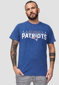 Royal blue cotton T-paita, jossa on valkoisella painettuna "New England Patriots" ja logon sekä tähtien design tekstin yläpuolella. Lyhyt hiha, pyöreä kaula-aukko.