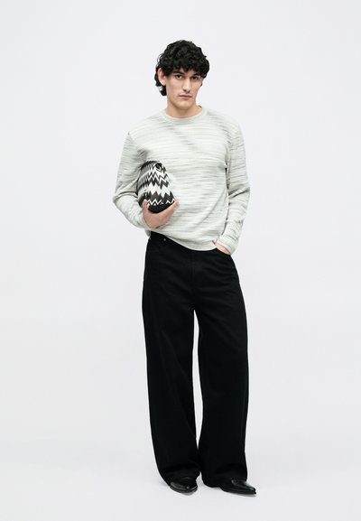 Gaiši pelēks teksturēts džemperis, melnas platajiem kājām bikses, rokās tur melnu un baltu zigzagveida raksta somiņu. Minimālistisks dizains, neitrālas krāsas.