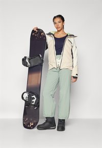 Snowboard s crnim i sivim vezovima, koju drži osoba u svijetlo bež izoliranoj jakni, tamnoplavoj majici i svijetlozelenim hlačama, nosi crne čizme.
