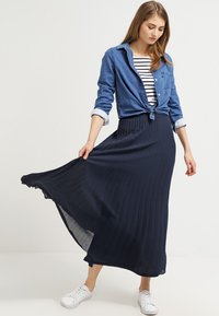 Marineblå plisseret maxiskirt parret med en knyttet denimskjorte over en stribet top; båret med hvide sneakers. Glat tekstur, svævende design.