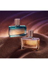 To bronse gudinneparfymeflasker fra Estee Lauder, en med blåtonet glass og en med klart glass, plassert på en glitrende bronsefarget sandbakgrunn.