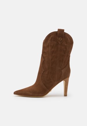 Botín de ante marrón con punta puntiaguda, costuras decorativas y un tacón apilado de 10 cm. Presenta una lengüeta en la parte posterior para facilitar el acceso.