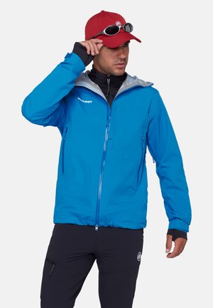 Man draagt een blauwe Mammut-jas, zwarte broek, rode pet met zonnebril erop, en past de pet aan met zijn rechterhand.