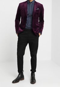 Man som bär en lila sammet blazer, blå randig skjorta, svarta byxor med uppvikta ben och svarta läderankelstövlar, står med händerna i fickorna.