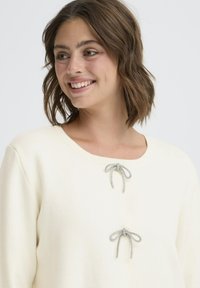 Kremni pulover z okroglo izreznico, ki na sprednji strani združuje dva srebrna dekorativna loka, izdelan iz mehkega materiala.