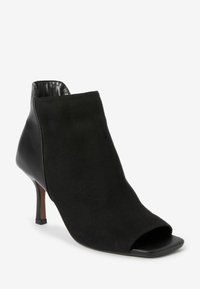 Stivale alla caviglia nero in suede con design open toe. Presenta un tacco lucido e slanciato e un pannello posteriore strutturato e liscio.