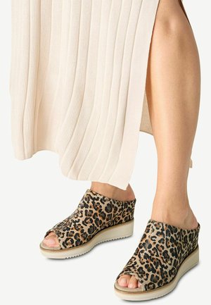 Kvinde iført beige ribstrikket nederdel og kilehælssandaler med leopardprint og åbne tæer, stående på en lys baggrund.