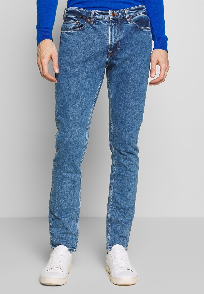 Homme portant un jean bleu, un haut à manches longues bleu et des baskets blanches, debout devant un fond clair uni.