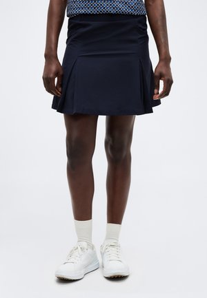 Persoon draagt een marineblauwe geplooide rok, witte sokken en witte sneakers, staand tegen een effen lichte achtergrond.