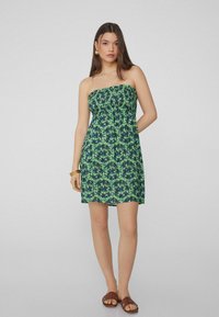 Trägerloses Kleid in Grün mit einem Palmenblattmuster. Rüschenoberteil und fließender Rock. Getragen mit braunen Sandalen und goldenen Accessoires.