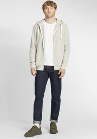 Lys grå zip-up hoodie med hvide snore, båret over en hvid t-shirt. Parret med mørkeblå jeans og olivengrønne sneakers.