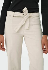 Buste portant un pantalon taille haute écru avec une ceinture nouée et un haut côtelé noir à manches longues, mains détendues de chaque côté.