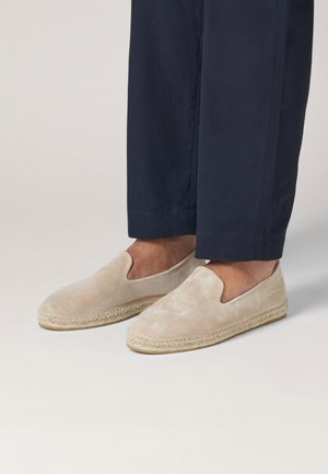 Beige suède instap espadrilles gedragen met marineblauwe cropped broek, met zichtbare details van de gevlochten juten zool en zonder zichtbare sokken.