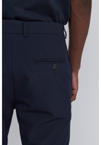 Navy blue trousers featuring a smooth fabric, slim fit, and a back patch pocket with a button closure.