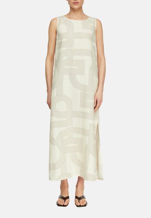Kremowa, bezrękawna sukienka maxi z beżowym geometrycznym wzorem linii, z bocznym rozcięciem, noszona z czarnymi sandałami z odkrytymi palcami na neutralnym tle.
