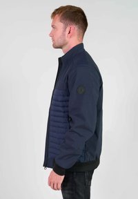 Marineblauwe jas met een gequiltte voorkant, geribde mouwen en een hoge kraag. Gladde textuur met een matte afwerking en een patchdetail op de mouw.