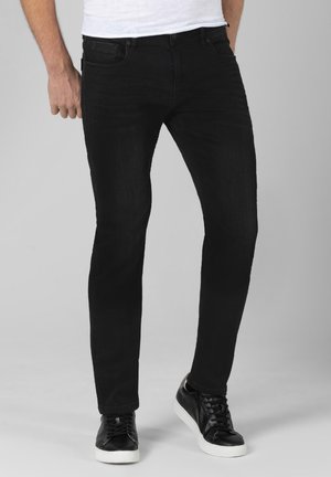 Mann trägt schwarze Slim-Fit-Jeans und schwarze Sneakers mit weißen Sohlen vor einem einfachen grauen Hintergrund.