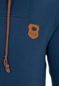 Sweatshirt bleu marine en tissu texturé, avec un patch en cuir marron et un cordon en leçon tressé. Design à col rond avec un minimum de détails.