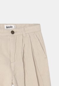 Beige korduroy kalhoty s texturovaným povrchem, které mají přední záhyby, boční kapsy a zapínání na knoflík s logem na pase.