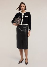 Svart och vit stickad cardigan med knappar, kombinerad med en svart läder pennkjol, hållande en metallisk handväska. Spetsiga lackskor.