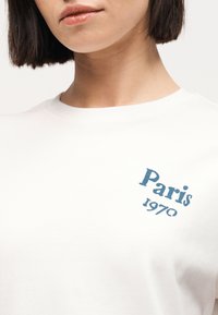 Γυναίκα που φοράει λευκό πουκάμισο με μπλε κείμενο "Paris 1970" στην πάνω αριστερή πλευρά του στήθους, ορατό από τον λαιμό μέχρι τη μέση του κορμού.